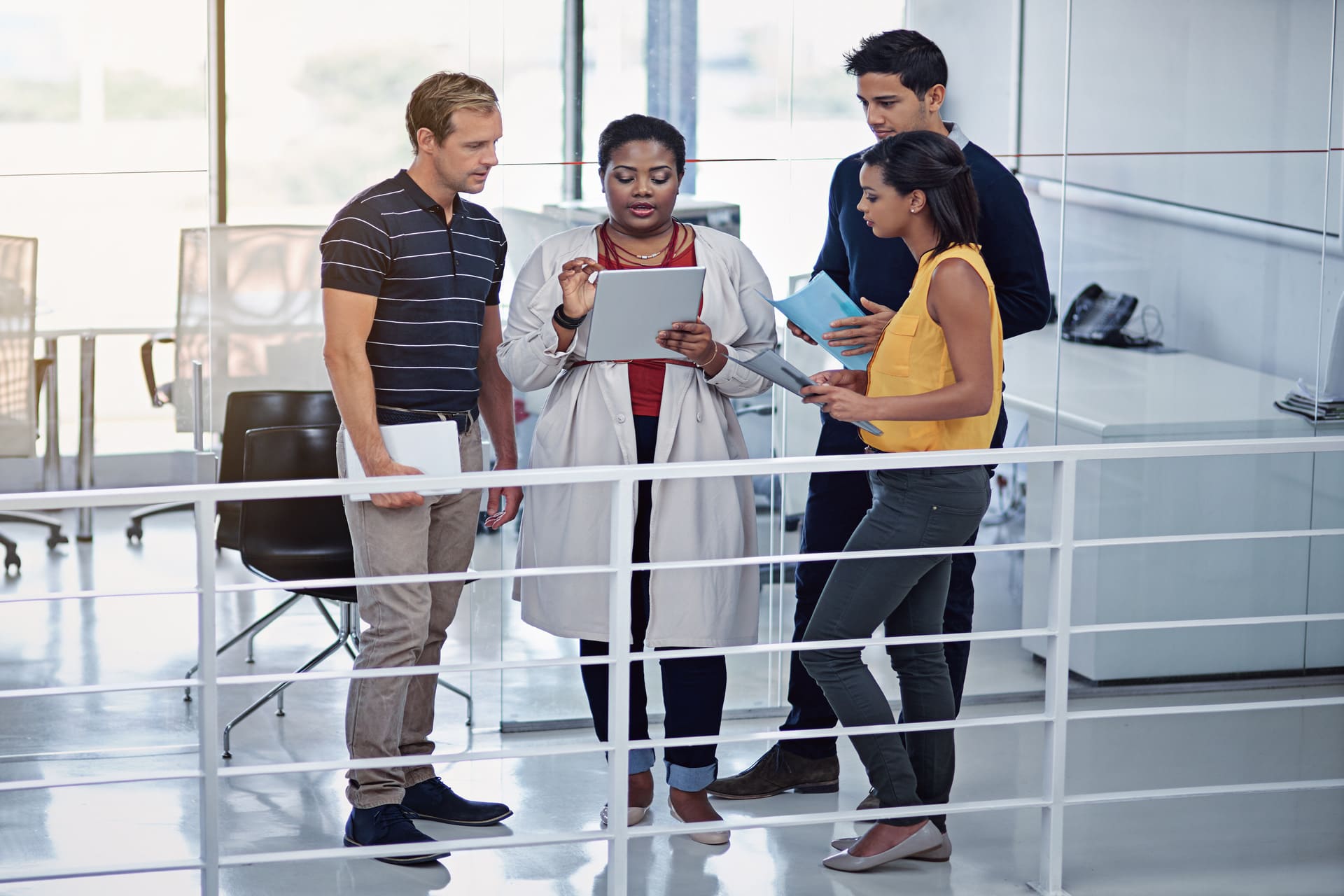 Four colleagues standing in a modern office discussing work while looking at digital tablets and documents