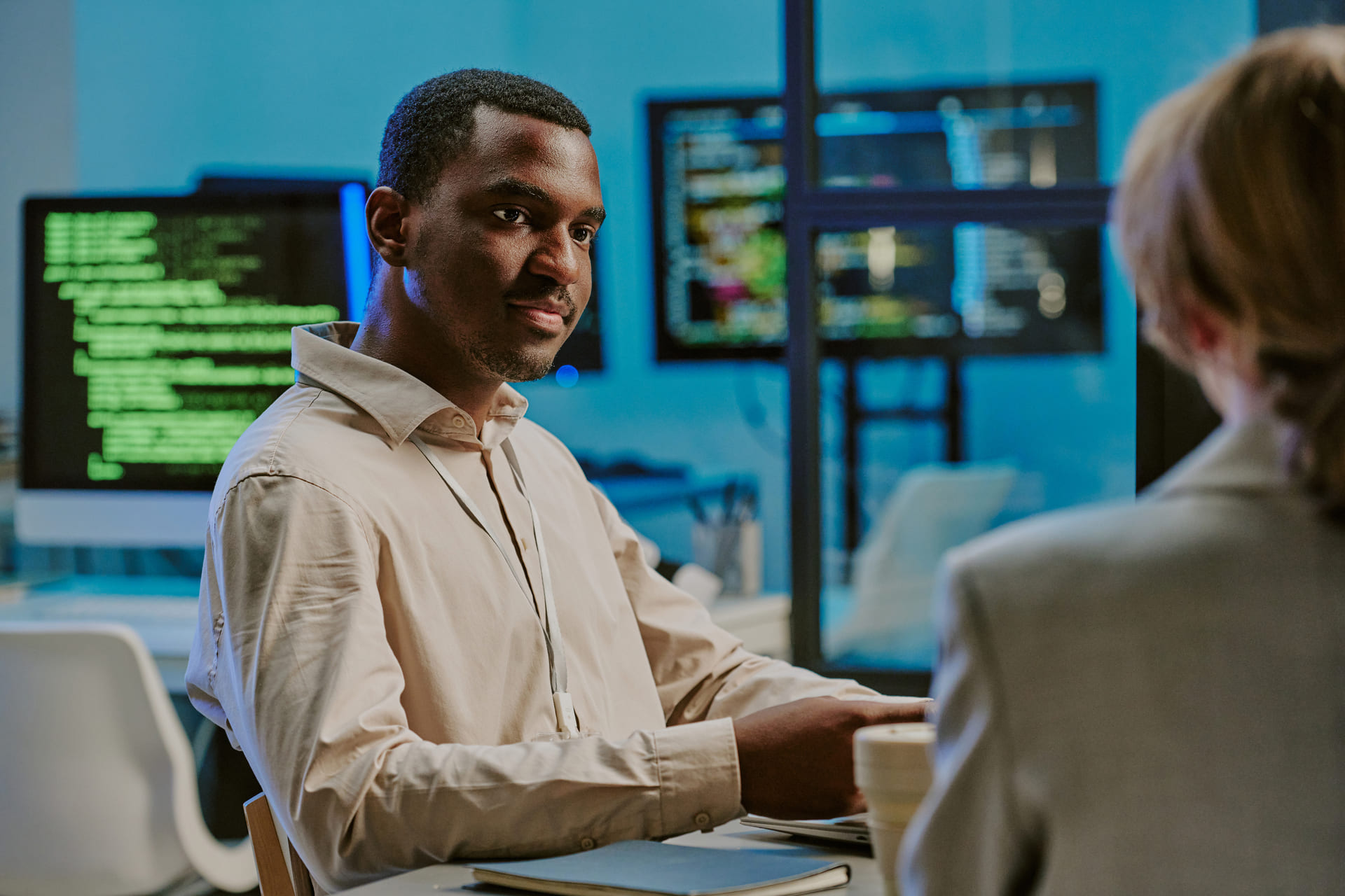 Man in a beige shirt speaking during a meeting in an IT office with computer code displayed on monitors in the background