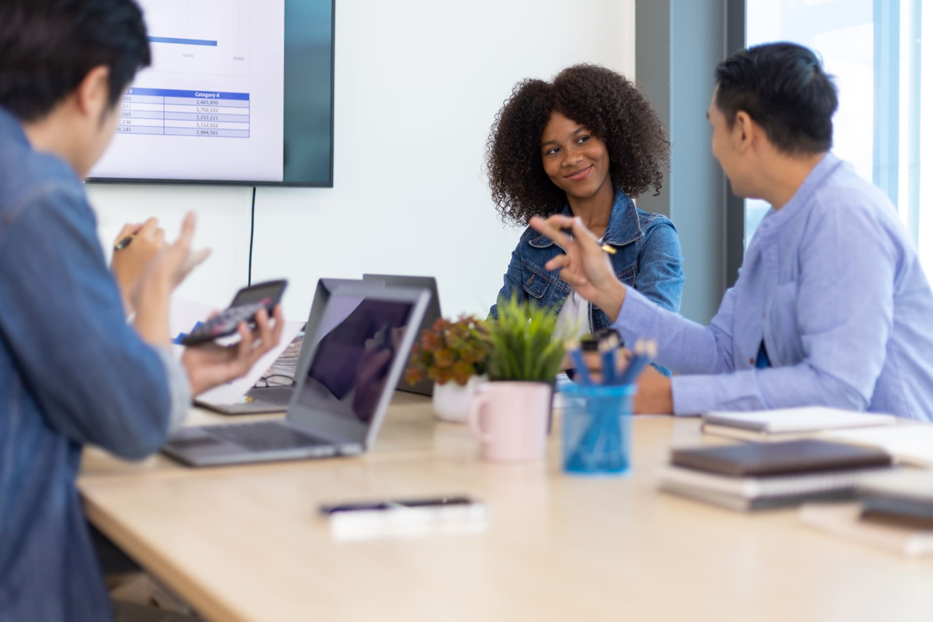 Three colleagues in a meeting room engaged in discussion, with laptops and a presentation screen showing data charts