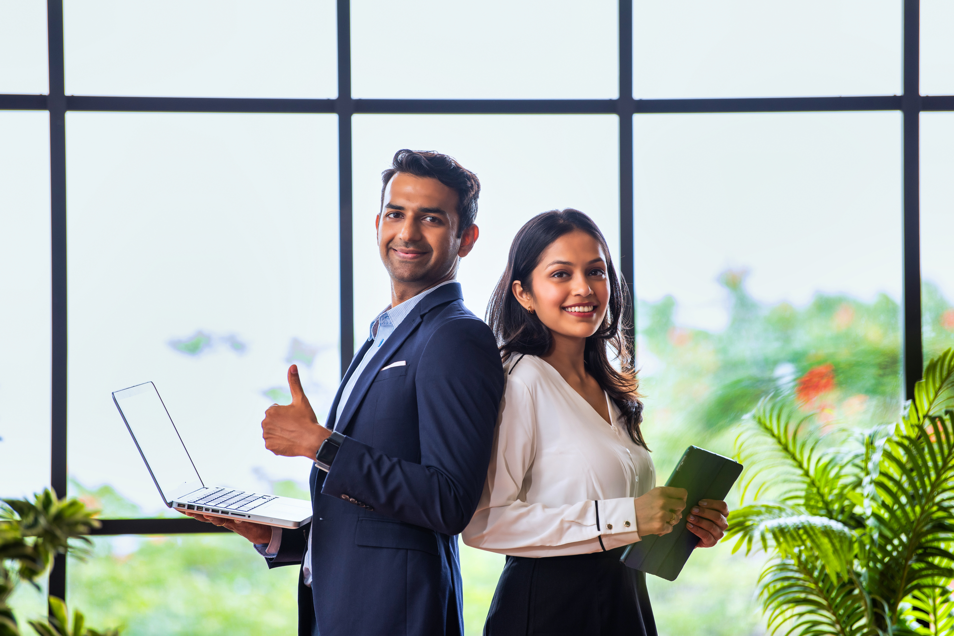 Two-smiling-business-professionals-standing-back-to-back-one-holding-a-laptop-and-giving-a-thumbs-up-the-other-holding-a-tablet-with-greenery-outside-large-windows