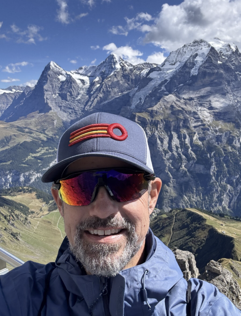 Man smiling at the camera in a blue cap and reflective sunglasses, with snow-capped mountains and green valleys in the background.